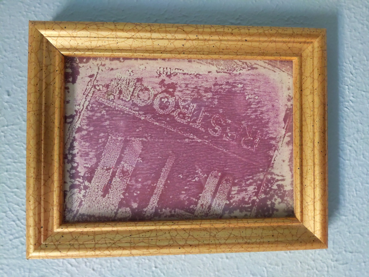 A bright pinkish-purple gel print monotype taken from an old restroom sign, framed in a gilt frame, and hung outside where the Sunrise PottyDoor will be installed.